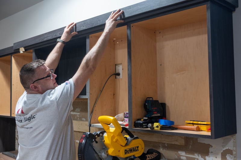 Installer fits a wood piece to the top of kitchen cabinets